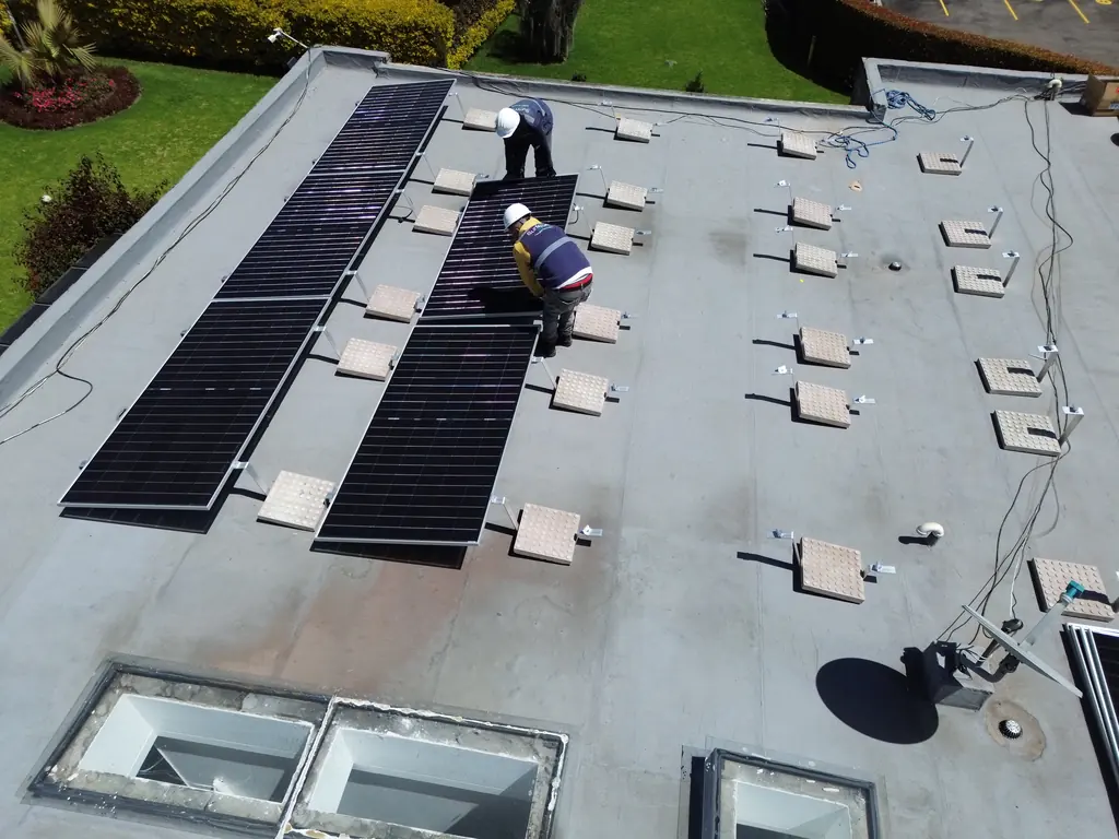 Vista cenital de dos filas de módulos solares instaladas sobre un techo plano con bases de lastre, mientras un técnico ajusta la estructura.