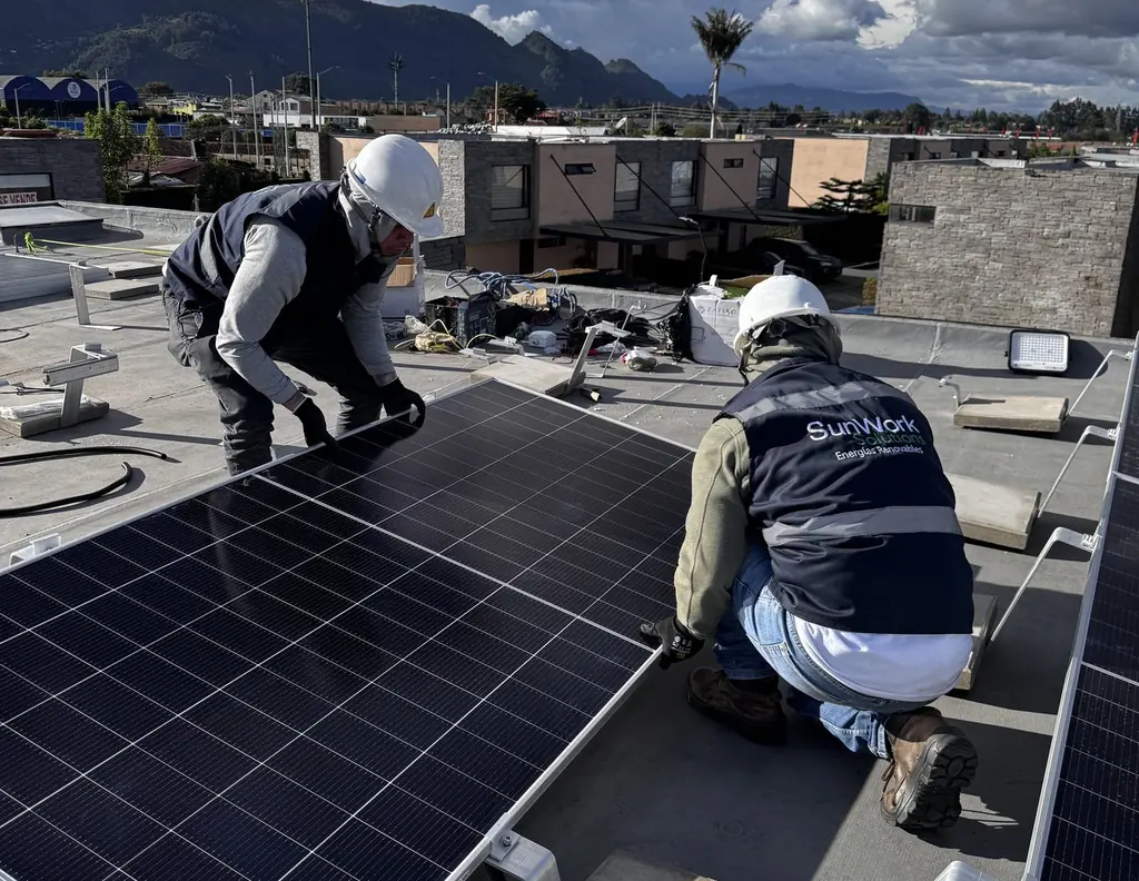 Dos técnicos de SunWork con casco y chaleco de la marca montan un módulo solar sobre un techo plano, con los cerros de la Sabana al fondo.